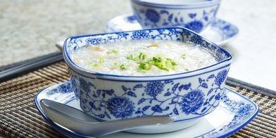 White porcelain bowl with blue flower pattern filled with congee garnished with green onions. The bowl is on a matching saucer with a spoon resting by it.