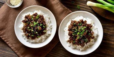 Two white bowls filled with white rice and topped with ground beef garnished with green onions and sesame seeds.